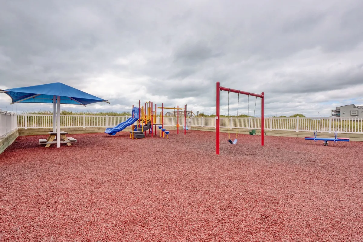 A colorful outdoor playground with swings and climbing equipment, set beneath a shade structure and accompanied by a picnic table. The area is lively, designed for children’s play and family gatherings.