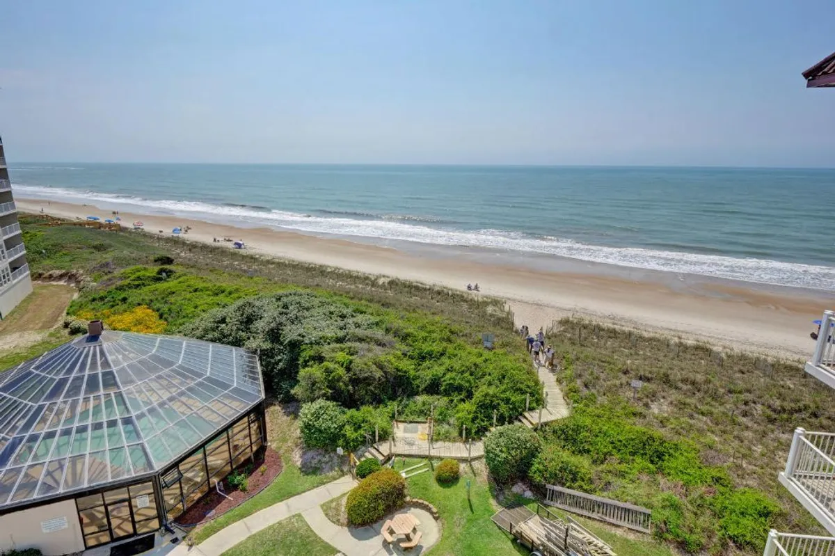 A tranquil view of the ocean and beach, bordered by a landscaped garden and a glass-enclosed seating area. The peaceful ambiance is accentuated by soft sunlight, perfect for enjoying quiet moments or sharing drinks while gazing at the Atlantic.