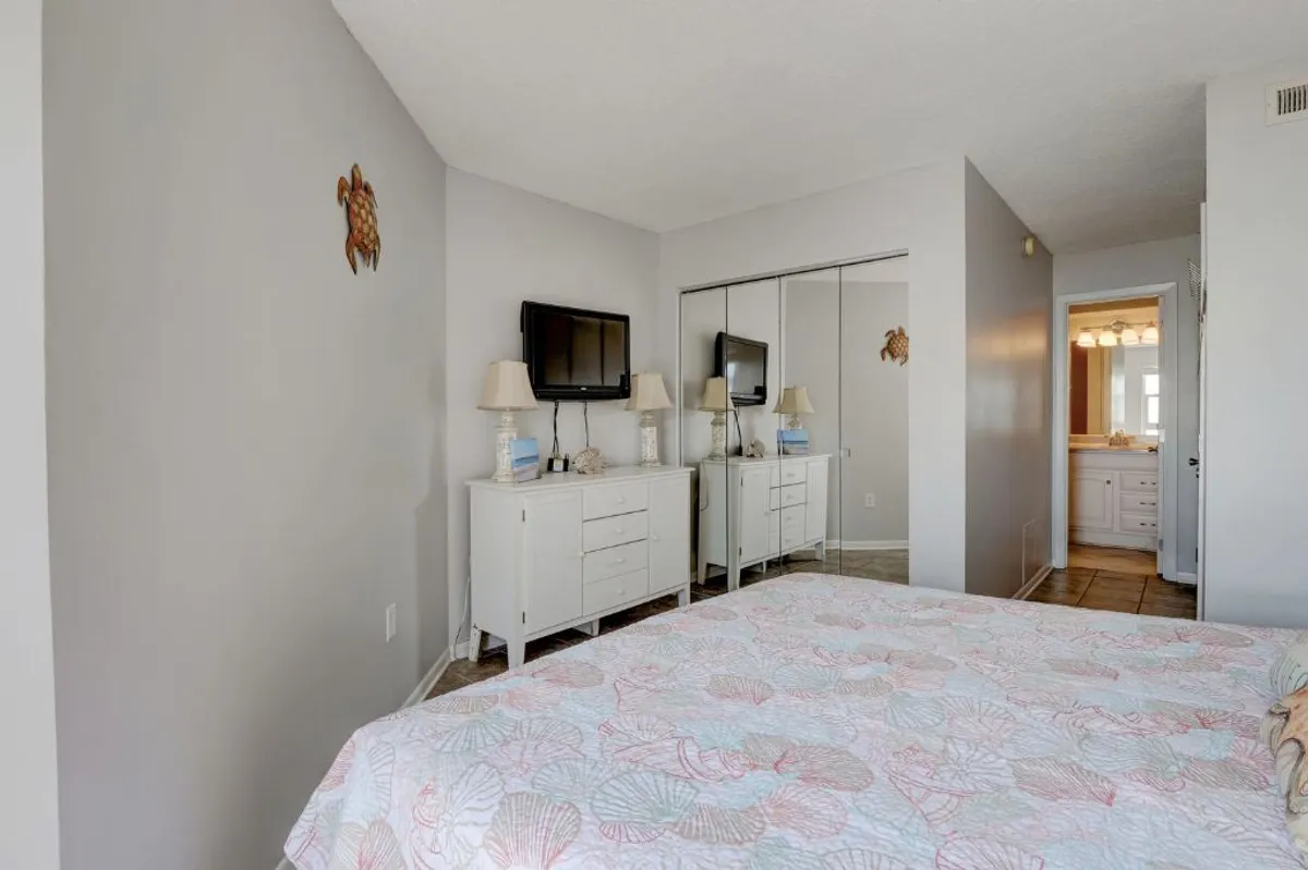 A serene master bedroom featuring a king-sized bed adorned with a patterned quilt, mirrored dresser, wall-mounted TV, and lamps on the nightstands. Soft natural light filters in, creating a peaceful and inviting atmosphere ideal for relaxation and privacy.