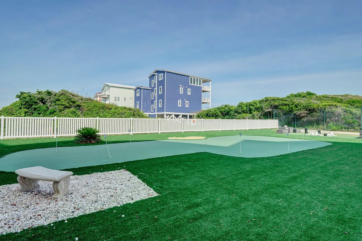 A landscaped outdoor area with an artificial putting green, decorative stone bench, and lush grass, inviting guests to practice golf or relax. The backdrop of a blue house adds a charming, leisurely vibe.