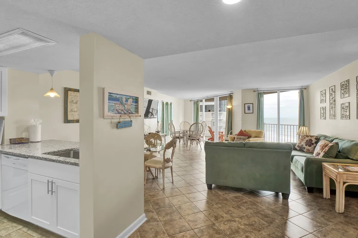 A broad perspective of the interconnected kitchen, dining, and living spaces, with a kitchen counter, comfortable seating, dining area, and large windows looking out to the ocean. The setup highlights ease of movement between spaces and opportunities for group interaction.