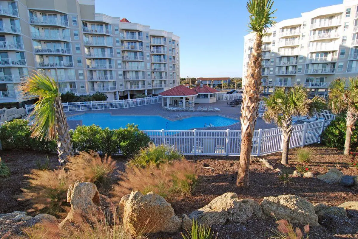 A sunlit outdoor pool surrounded by palm trees and lounge chairs, with condominium buildings visible in the background. The scene captures swimmers in the pool and people relaxing on the pool deck, evoking a sense of leisure and vacation.