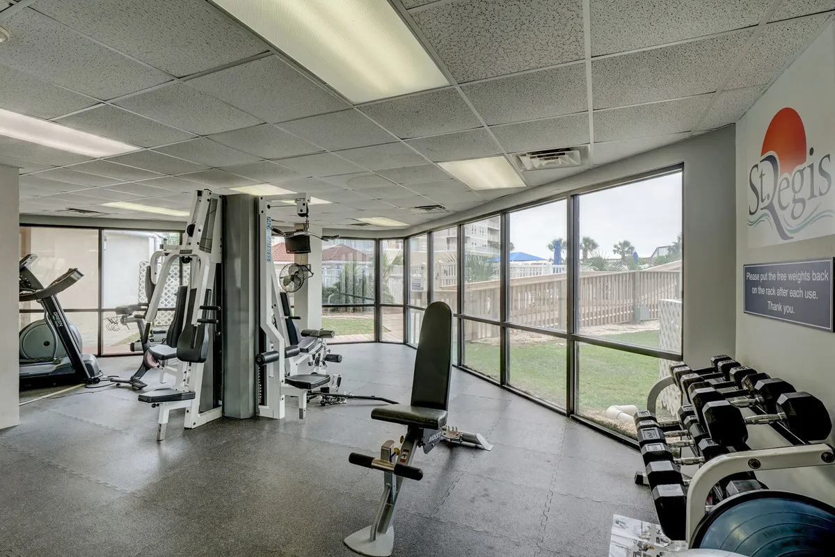 A bright, modern fitness center featuring a variety of exercise machines, weight racks, and large windows that let in natural light. The space is designed for workouts and group fitness activities, highlighting a well-equipped health and wellness facility.