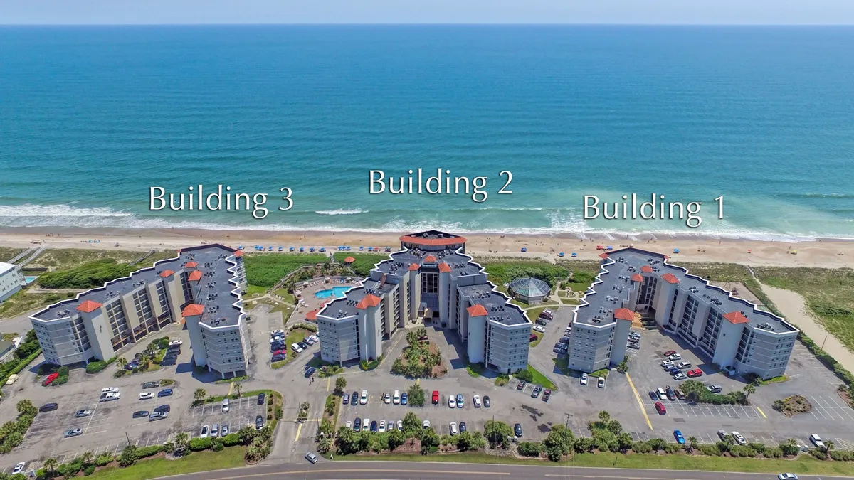 This exterior image shows three condo buildings with a sparkling swimming pool in the foreground and sweeping views of the ocean in the background. There are guests visible swimming, sunbathing, and preparing for a day of beach walks, encapsulating the lively yet relaxed atmosphere of a resort by the sea.