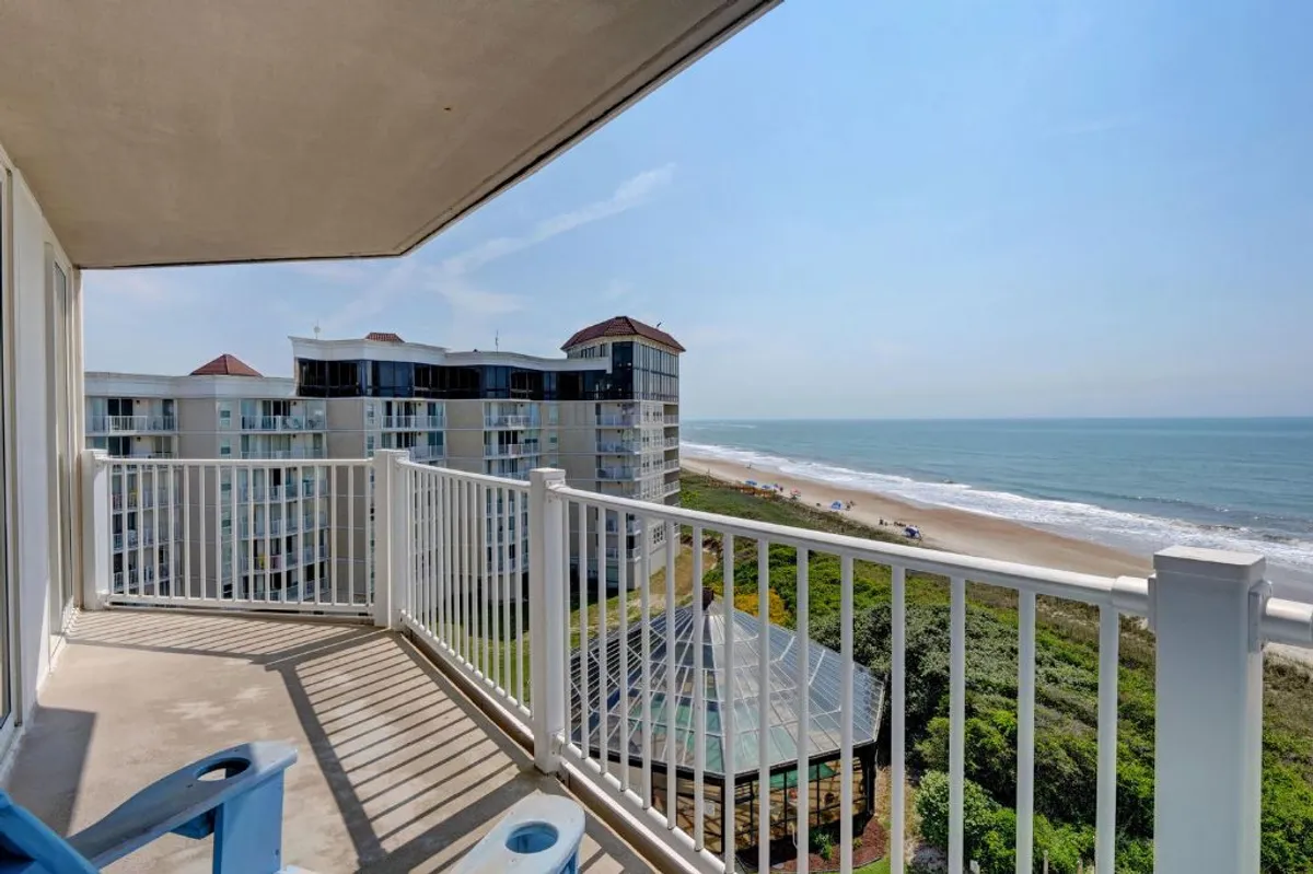 The image showcases a serene beachfront view from a private balcony, with the expansive ocean stretching to the horizon and soft sunlight glinting on the waves. A sleek balcony railing frames the scene, inviting guests to relax and take in the tranquil sunrise or unwind under a starlit sky. Nearby buildings hint at a friendly coastal community, while the wide sandy beach below promises endless opportunities for strolls and gatherings.
