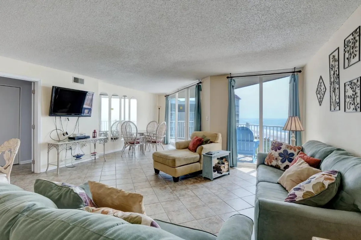 This image showcases a spacious, sunlit living room with comfortable seating, large windows framing sweeping ocean views, and an adjacent dining area with wicker chairs. Decorative pillows and a cozy, casual atmosphere invite relaxation and togetherness.