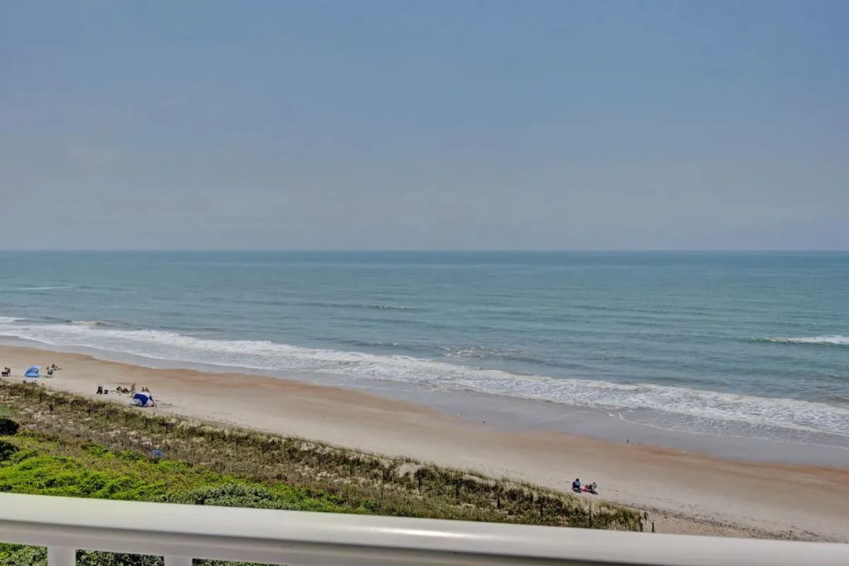 A wide view of the beach with the ocean stretching to the horizon. People are seen enjoying sunbathing, swimming, and strolling under a broad blue sky, evoking the sense of freedom, relaxation, and adventure available just steps from the condo.