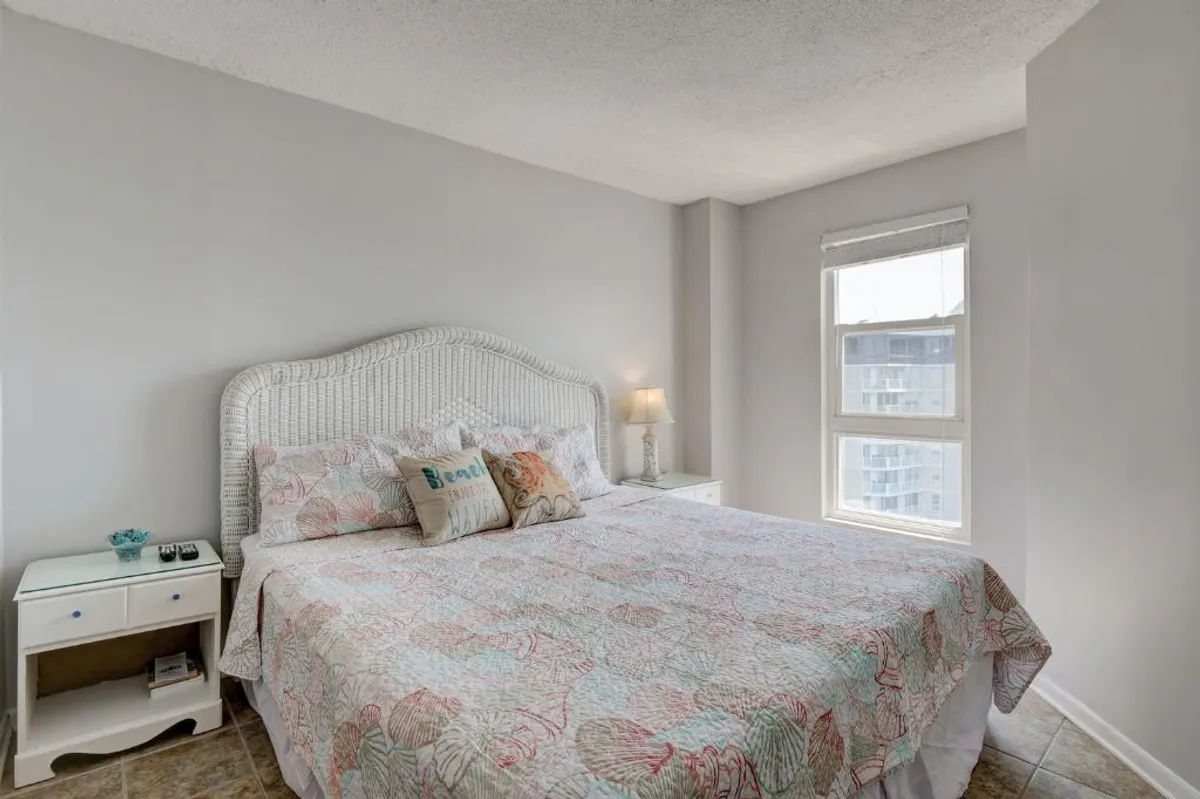 A cozy bedroom with a colorful quilt on the bed, a nightstand with a lamp, and a window letting in natural light—creating an inviting space perfect for winding down or enjoying a morning coffee.